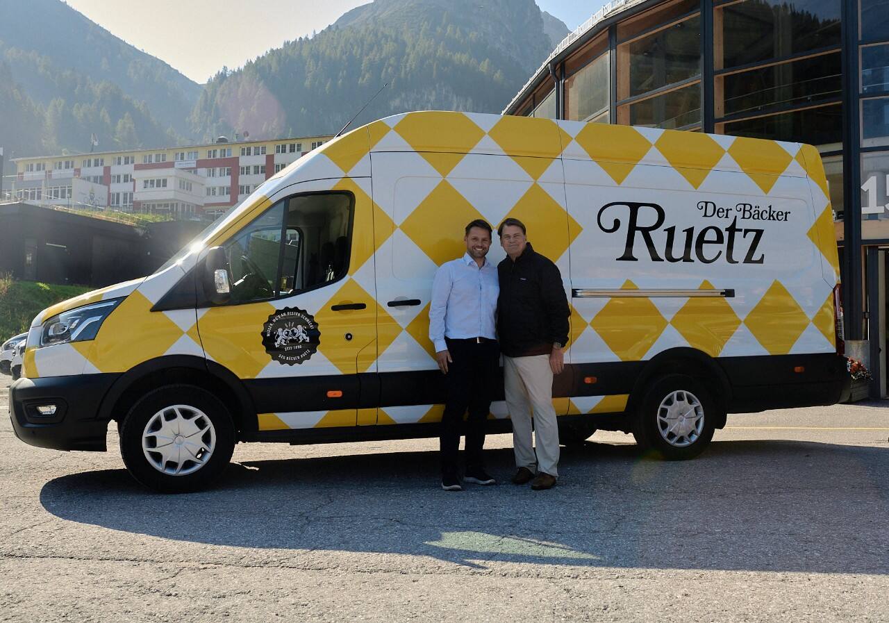 Two men stand in front of a yellow and white checkered truck with the words Der Backer Ruetz on the side.